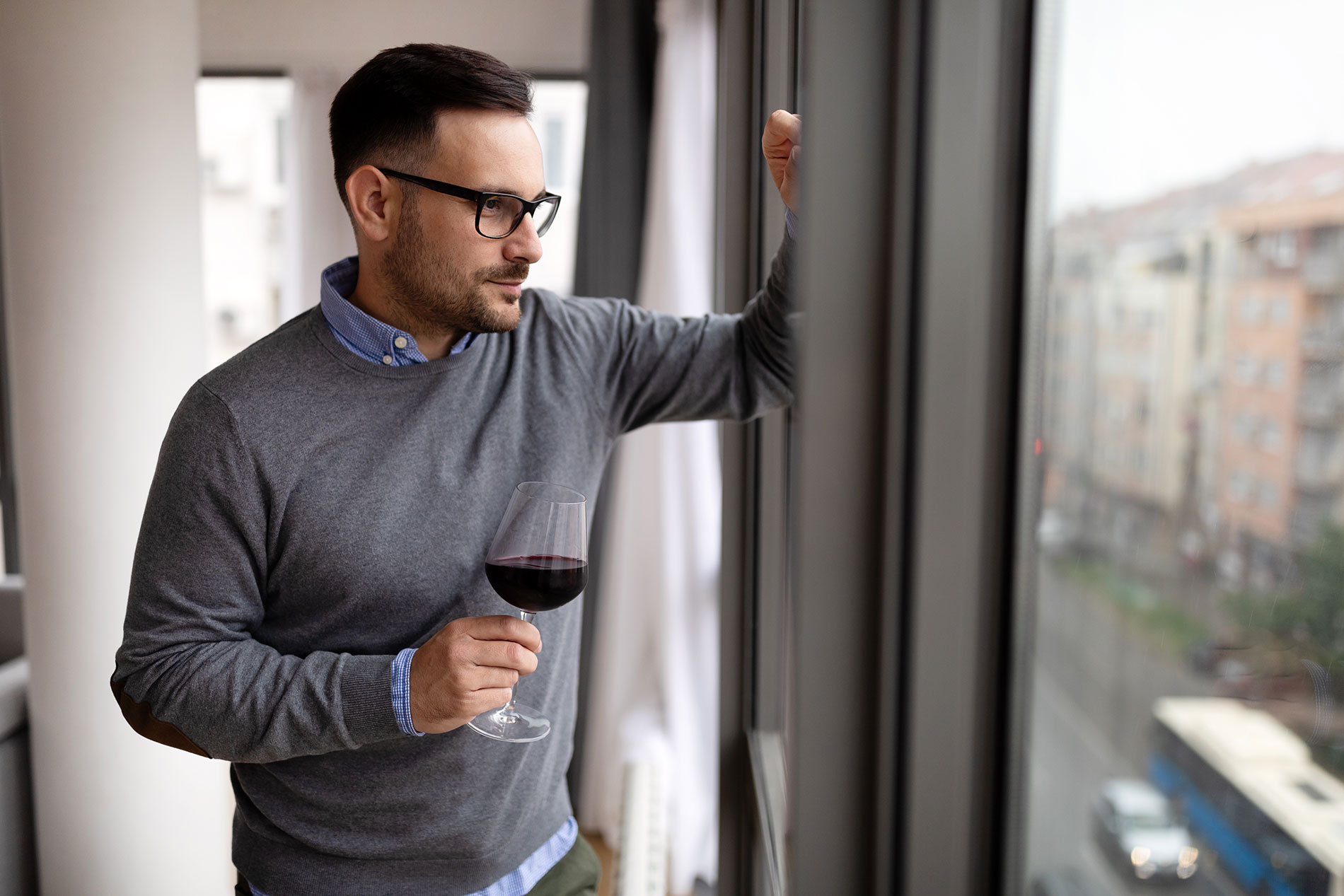 Ein junger Mann genießt ein Glas Rotwein am Fenster: Alkohol hat vielfältige Wirkungen auf Körper und Psyche.